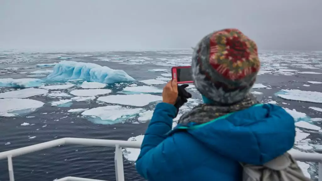 South Spitsbergen traveler in knit hat & blue jacket stands on ship deck & photographs blue iceberg on a misty day.