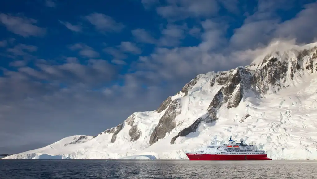 Small polar expedition ship with red hull & white upper decks sits in glassy sea beside tall, snowy cliffs in Antarctica.