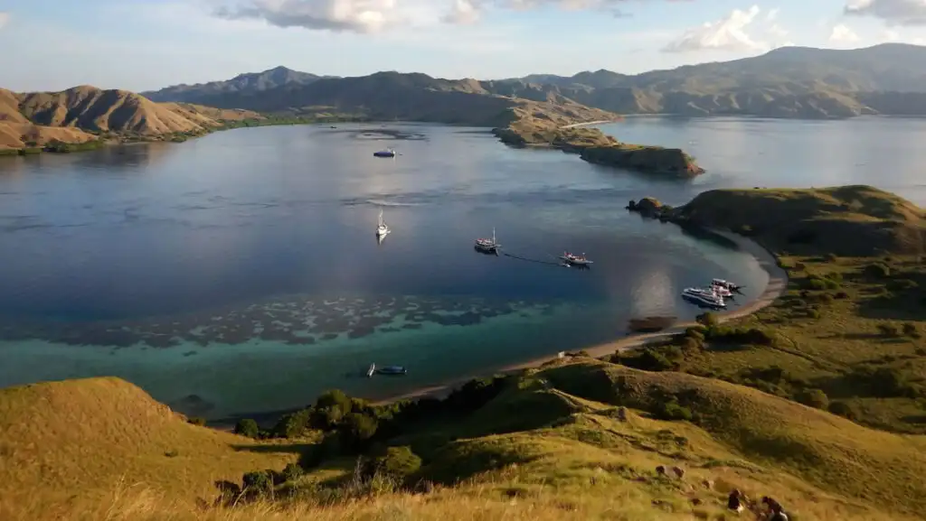 Aerial view of Indonesian archipelago with blue & turquoise sea, hills draped in green & gold and small ships sitting at anchor.