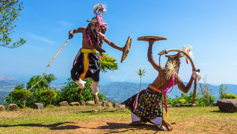 2 men in colorful native garb on a mountaintop do a demonstration fight dance with wood stalks & shields in the Indonesian sun.