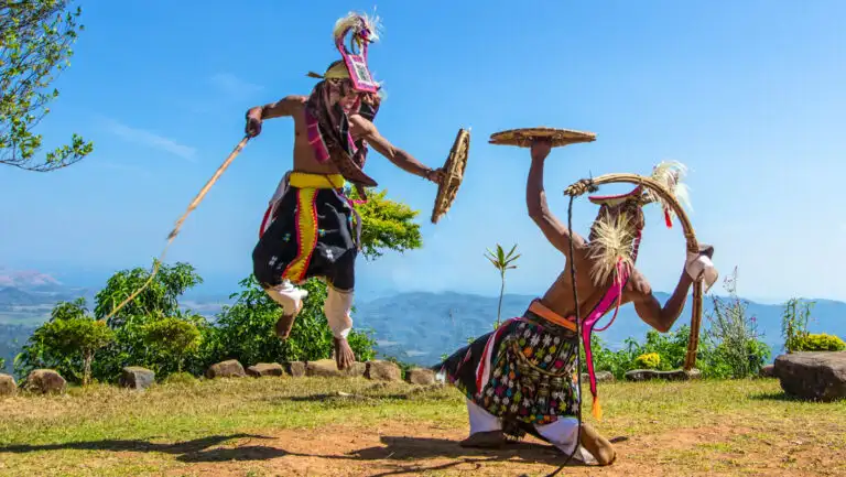 2 men in colorful native garb on a mountaintop do a demonstration fight dance with wood stalks & shields in the Indonesian sun.