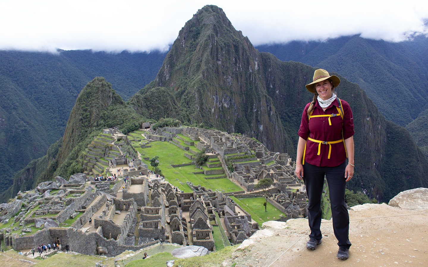 A female traveler in a tan safari hat and red shirt stands on a hillside with Machu Picchu below her