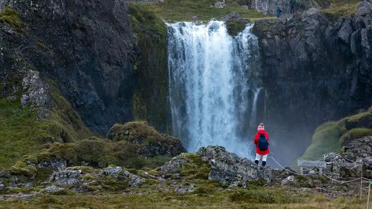 hiker in red standing near beautiful Waterfall with black rocks and green landscapes