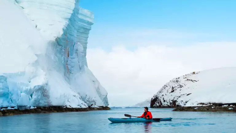 Man in bright red jacket paddles blue kayak in calm sea past towering glacier of snow & ice on a blue-sky day in Antarctica.