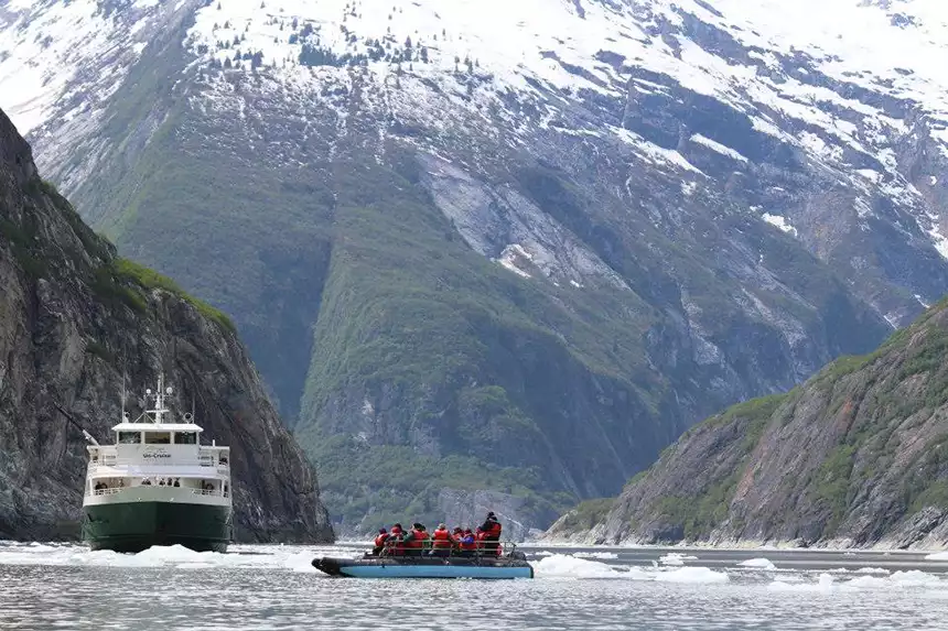 A mountainous fjord is the setting for a small ship seen behind a Zodiac with a group of travelers in it