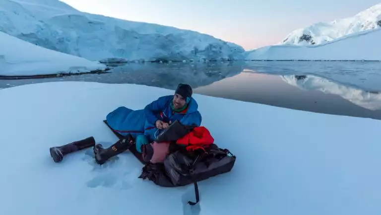 Man lays in down sleeping bag on untouched snowfield by glassy water & snowy mountains at dusk on the Antarctica Active cruise.