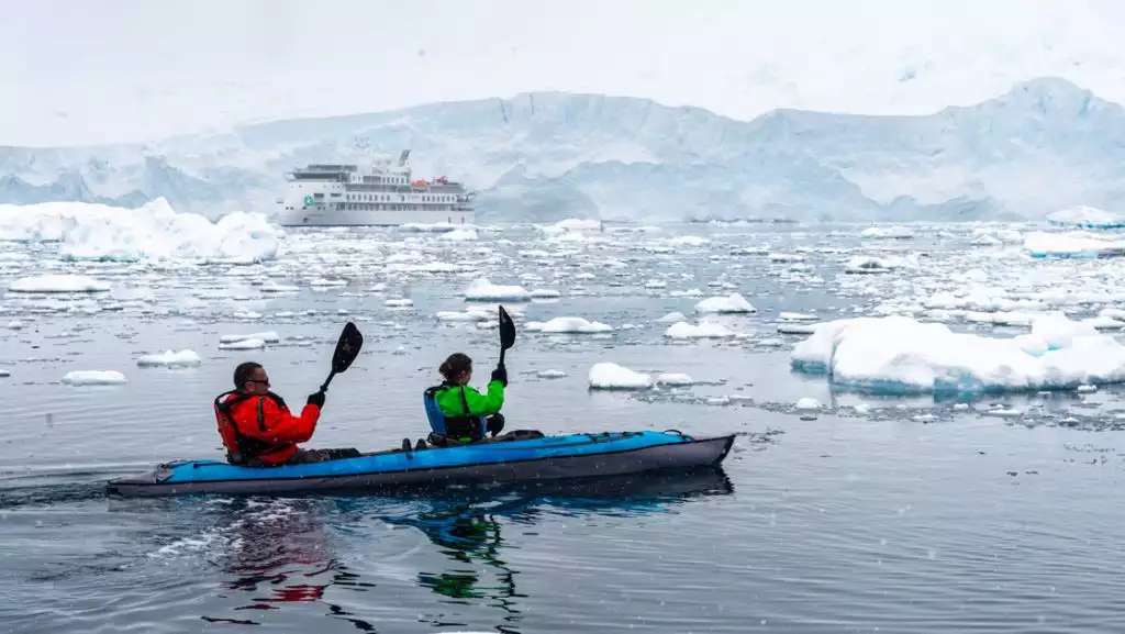 Tandem kayakers in red & green jackets paddle an inflatable blue kayak among icy waters by white expedition ship.