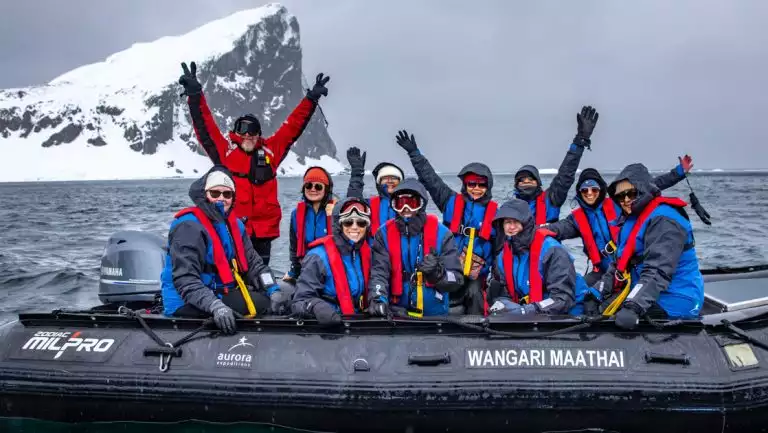 Black Zodiac boat with Antarctica Active cruise guests in blue jackets & red PFDs waving near a cliff on a cloudy day.