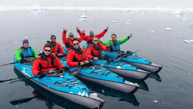 4 tandem inflatable teal kayaks sit side-by-side in calm sea as their Antarctica Complete cruise guests smile & wave.