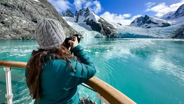 Woman in teal down coat & knitted hat holds camera over deck railing to photograph a blue glacier & snowy peaks in Antarctica.