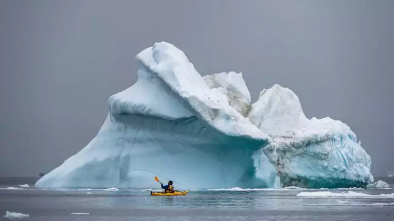 Kayaker passes by large iceberg floating in the middle of the water in a yellow kayak with winter gear