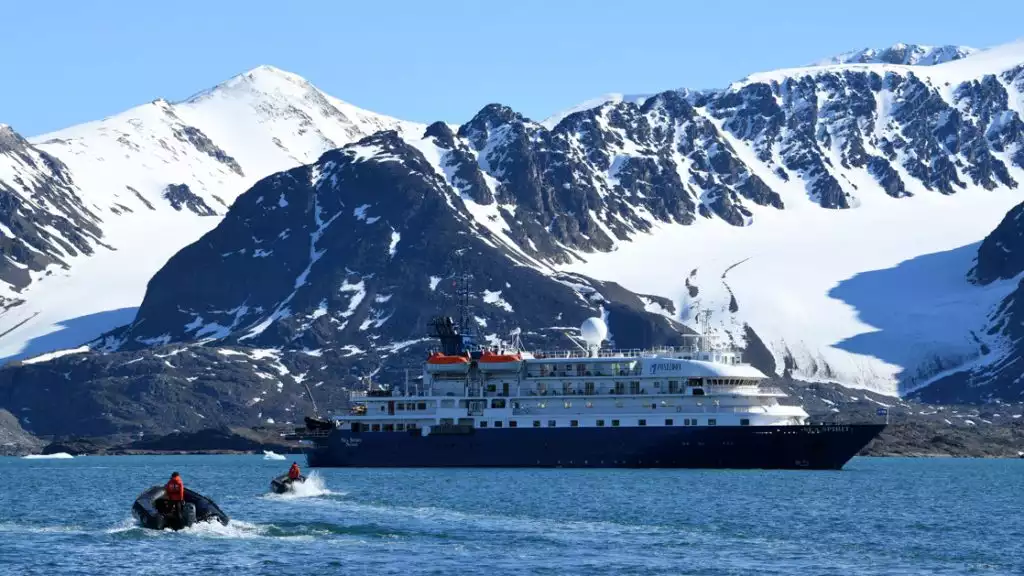 A small cruise ship sits in front of a mountainness landscape with two zodiacs exploring around the blue waters