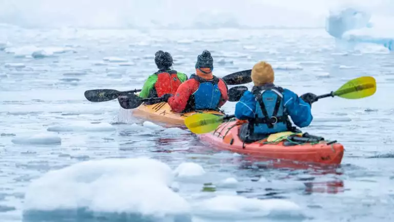 3 kayakers on a Falklands, South Georgia & Antarctic Peninsula cruise paddle red & orange boats among iceberg bits in a snowstorm.