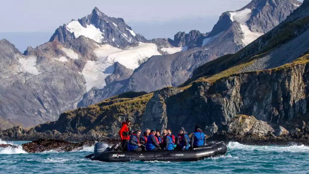 Zodiac dinghy with Falklands, South Georgia & Antarctic Peninsula cruise guests motors past dramatic peaks of rock, snow & flora.