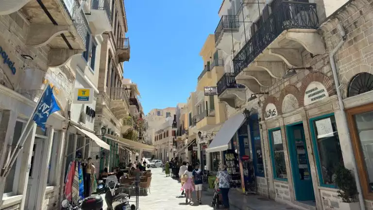 Greece and Turkey cruise travelers walk on white stone street past storefronts & under decks & awnings on a sunny day.