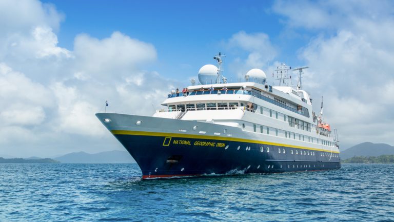 National Geographic Orion small ship with dark blue hull & white upper decks sits at sea in sunshine as guests wave from bow.