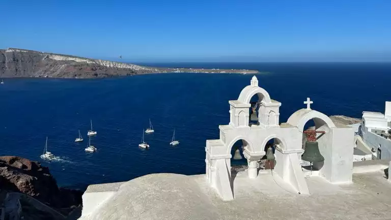 Whitewashed bell towers look out over deep blue Aegean Sea with small white sailboats & volcanic land behind, in Santorini.