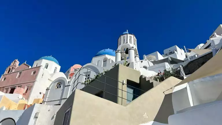 Sugar cube buildings in white, beige & pink with blue dome tops sit on hillside under deep blue sky in Santorini, Greece.