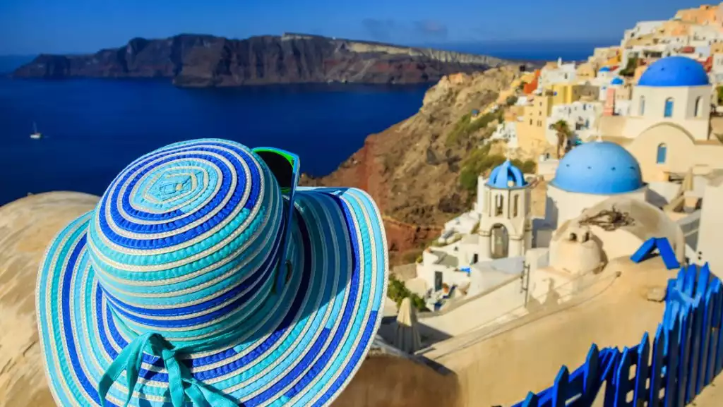Traveler in wide-brimmed blue & turquoise sun hat looks out over Santorini mountaintop town with blue domes & beige buildings.