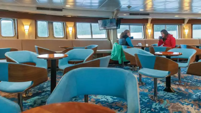 2 women sit among tables & chairs in window-lined lounge aboard National Geographic Sea Bird expedition ship.