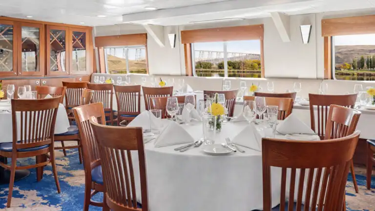 Dining room on National Geographic Sea Lion cruise ship with 2- & 4-top tables set in white linens surrounded by wooden chairs.