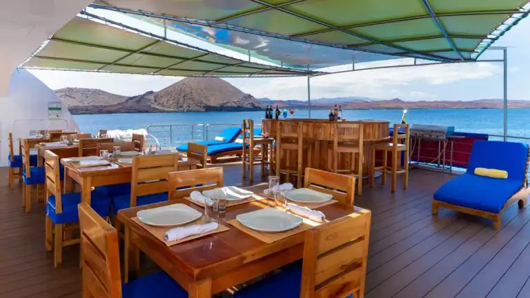 Outdoor dining area with wooden chair and blue cushions on small cruise ship overlooking blue waters and rocky mountains