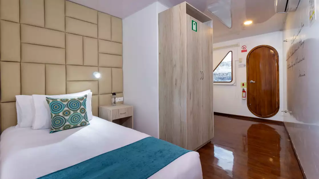 Single cabin on Petrel boat in Galapagos with twin bed with white sheets & padded gold headboard by white wall & armoire.