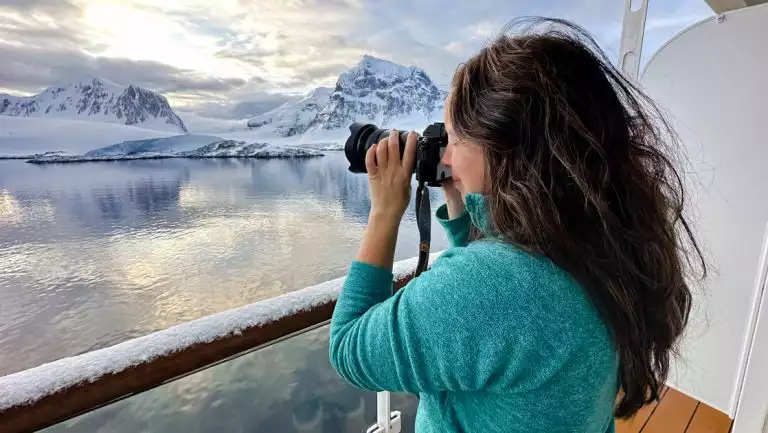 Woman in teal sweater stands on ship deck & photographs a white polar landscape in Antarctica during the Ross Sea Odyssey.