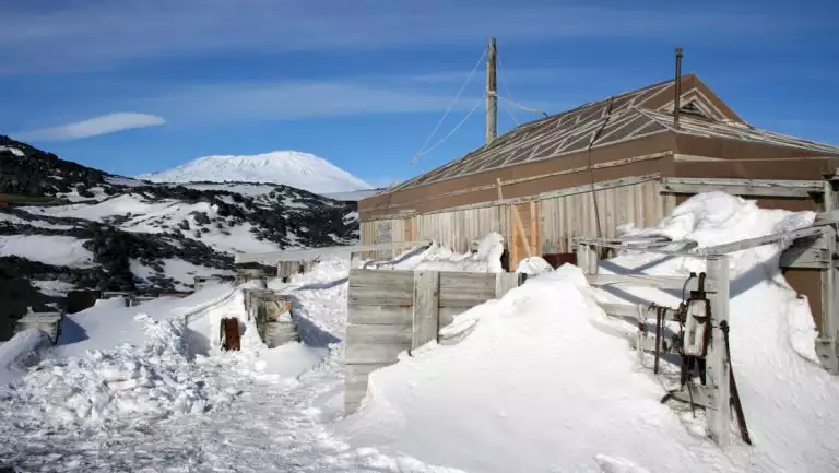 Rustic, historic wooden Shackleton hut sits among snow & ice by dark mountain under blue skies in Antarctica.