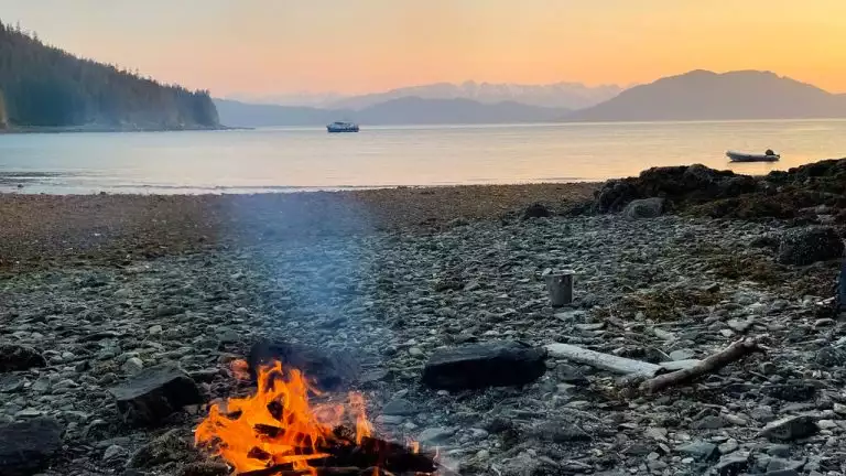 Individual seated by a campfire on the beach, gazing out at the tranquil sikumi-alaskan cruise