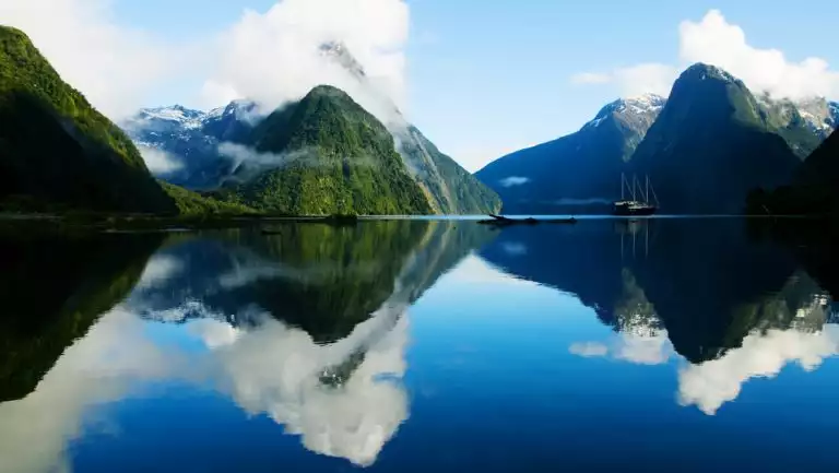 Fiordland, NZ, with small sailing ship sitting in glassy water that reflects the bright green gumdrop mountains all around.