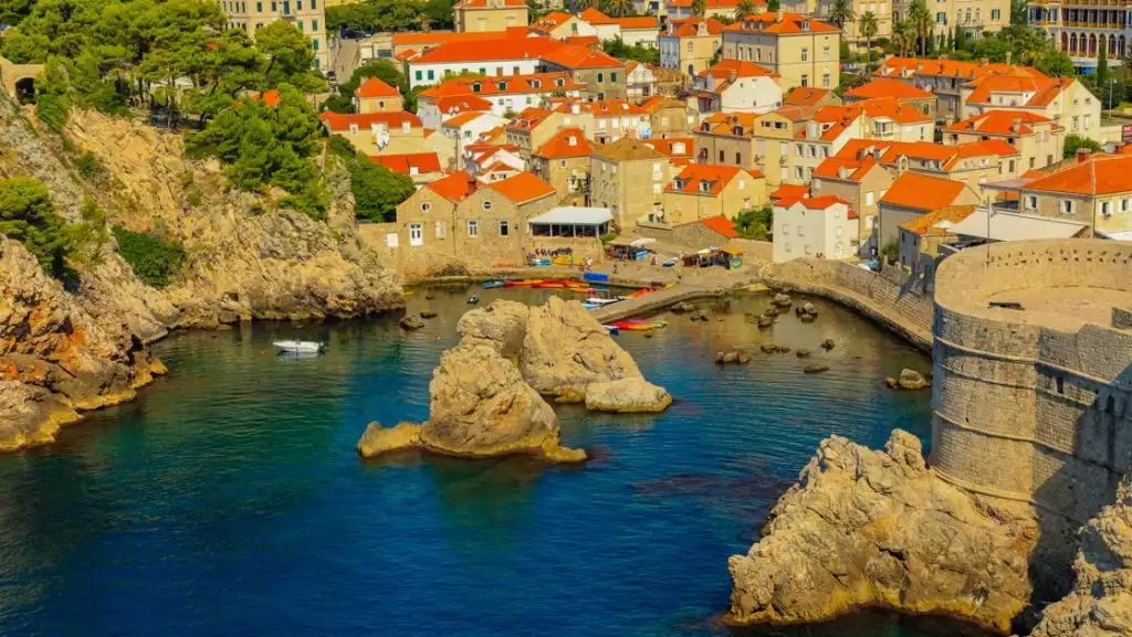 Blue-green Aegean Sea touches Dubrovnik Croatia's cove with kayakers & sun seekers below terracotta roofs.