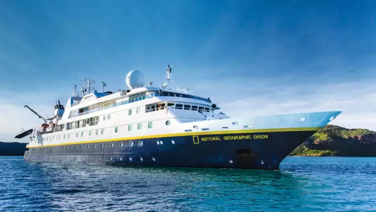 National Geographic Orion small ship with dark blue hull, yellow accents & white upper decks sits in turquoise sea in the sun.