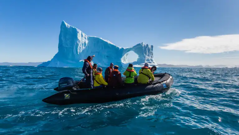Black Zodiac boat with Greenland travelers in colorful jackets cruises past a large iceberg with a circle cut out.
