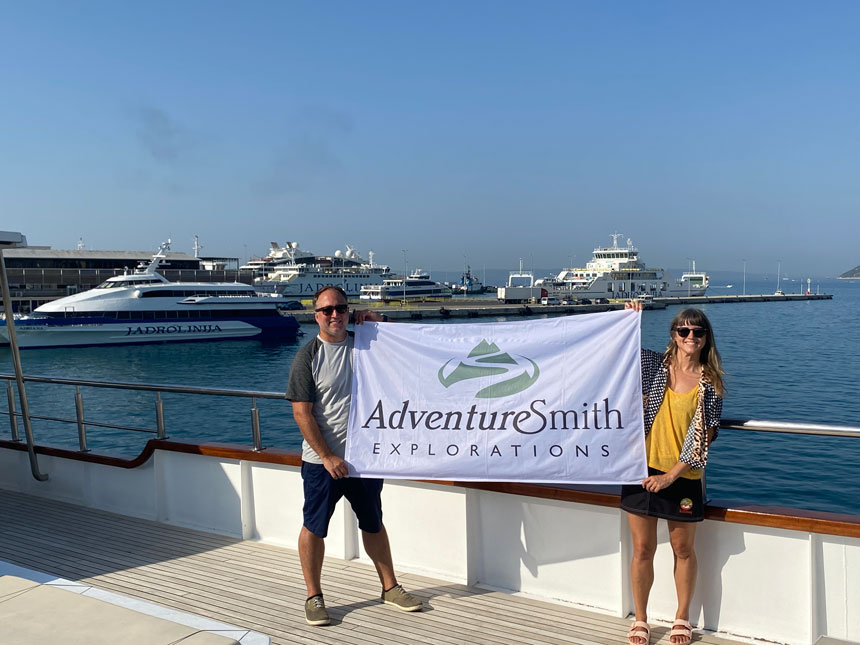 Man & woman hold a white & green AdventureSmith Explorations flag on open-air deck of M/S Aurora in Croatia.