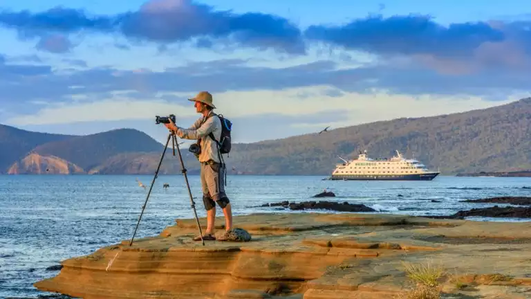 A photographer at sunset, on the Rocky Shoreline, with the ship National Geographic Endeavor II in the background, Santiago Island, Galapagos Islands, Galapagos National Park, Ecuador.