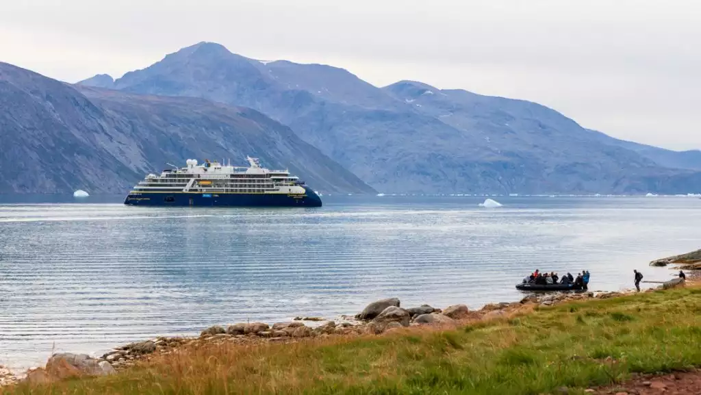 Small ship with dark blue hull & white upper decks sits offshore from grassy coast where Zodiac of Greenland travelers are arriving.