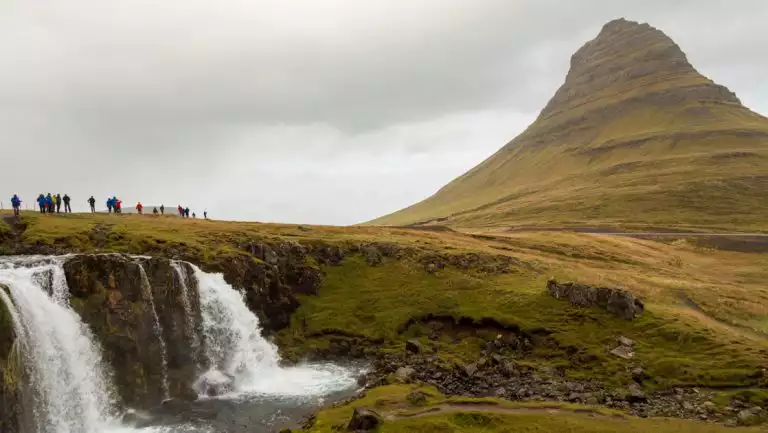 South Greenland Exploration cruise travelers stand atop the Kirkjufellsfoss waterfall near a grassy peak on a cloudy day in Iceland.