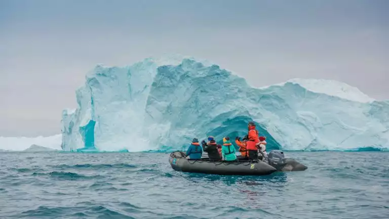 Zodiac boat with south Greenland Exploration cruise guests in colorful coats sits in front of large blue iceberg in calm sea.