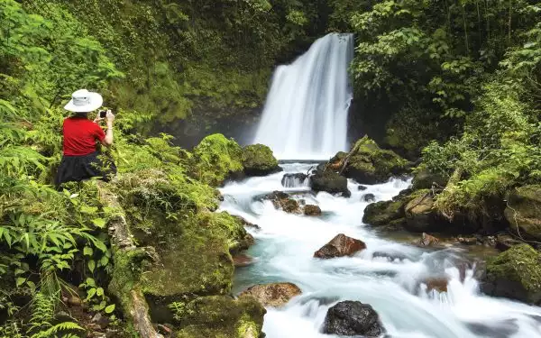 A female traveler in a red shirt and white wide brimmed hat kneels in lush green forest photographing a waterfall in the jungle of Costa Rica.