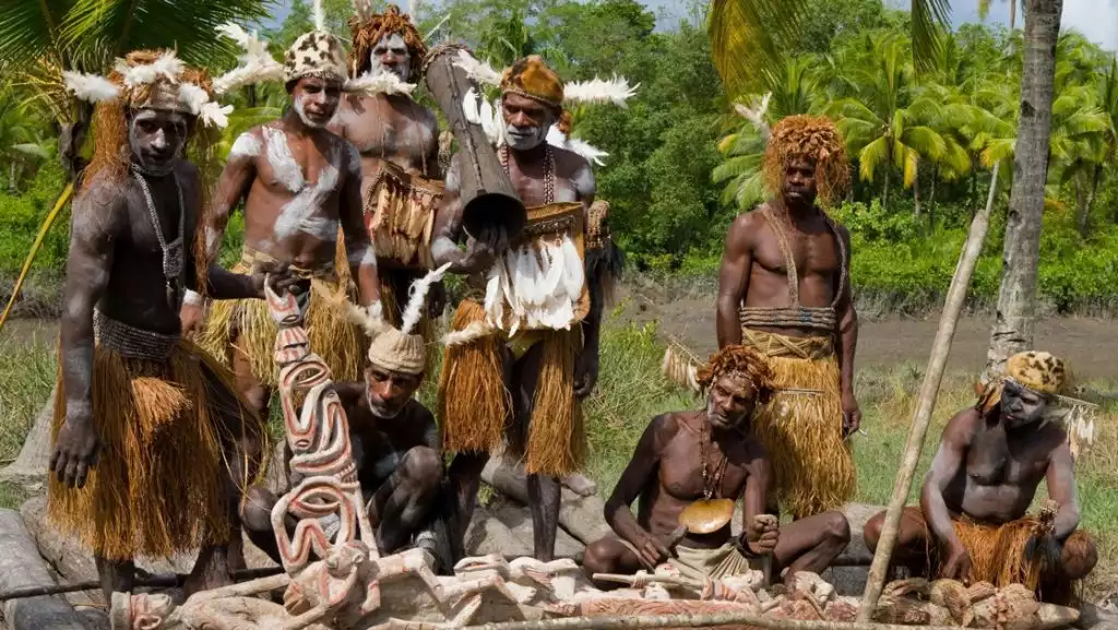 Native men of the Asmat Tribe of West Papua gather around a painted wooden carving with jungle behind them on a sunny day.