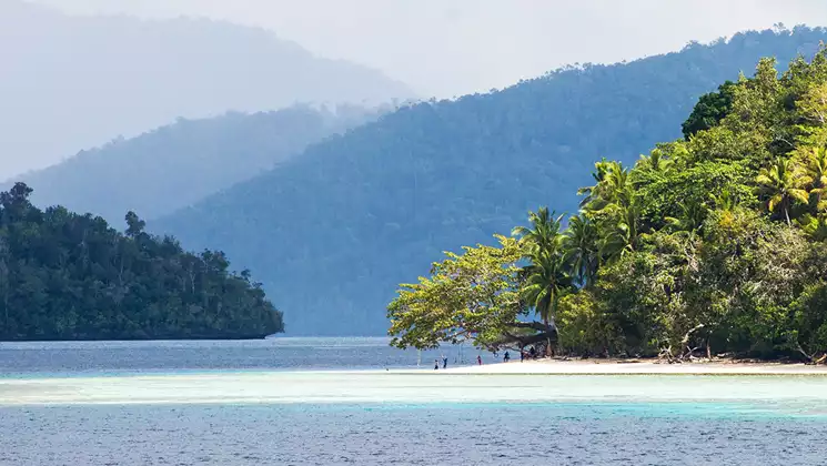 Small group of people play on a white-sand beach under palm trees by turquoise water backed by lush mountains in Indonesia.