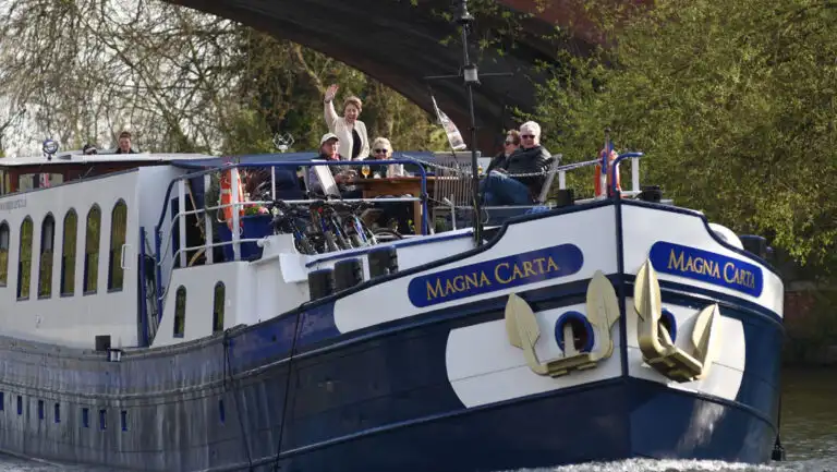 Classic England River Thames barge cruise guests wave from the bow deck as their small blue & white ship cruises under a bridge.