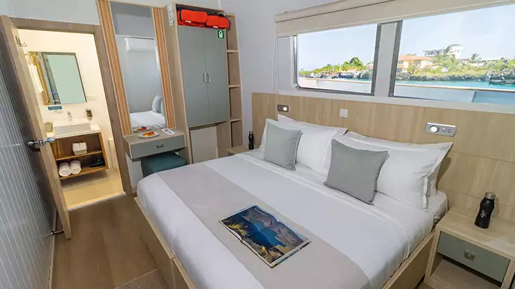 View of the Main Deck cabin aboard Galapagos Angel showing a King bed with a window behind it and a bathroom seen with door open