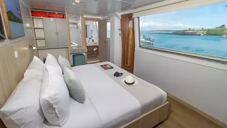 A suite aboard the Galapagos Angel yacht with a king size bed facing a large panoramic window showing the bright blue ocean and green shoreline outside