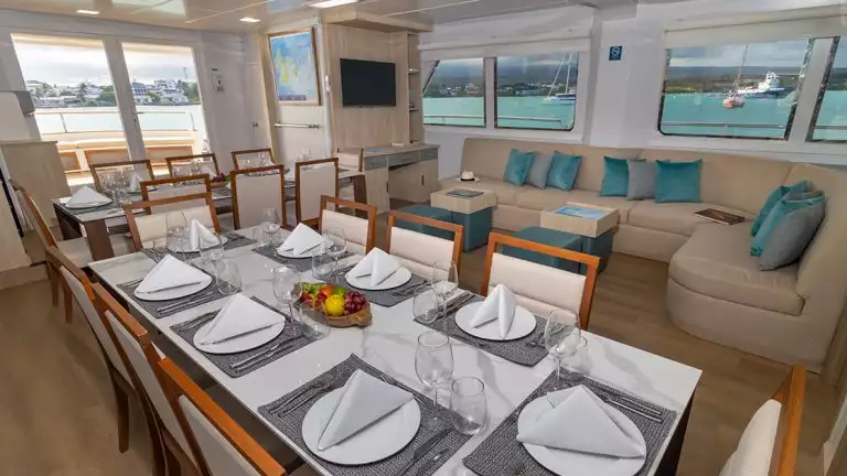 The combined lounge and dining area aboard Galapagos Angel with tan couch seating with blue pillows and windows lining the room