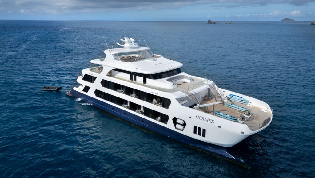 Aerial view of Hermes Galapagos luxury ship with 4 guest decks in white & blue with teak decking, sitting in calm sea.