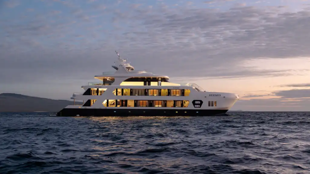 Hermes yacht in Galapagos at dusk with 4 white upper decks, 2 interior lit & 2 al fresco, and dark blue waterline deck.
