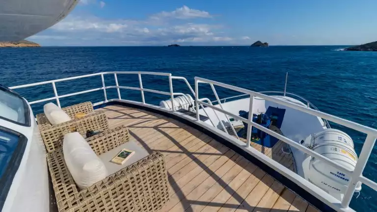 Forward-facing small teak deck with 2 padded chairs facing blue sea & small volcanic islands on Monserrat boat in Galapagos.