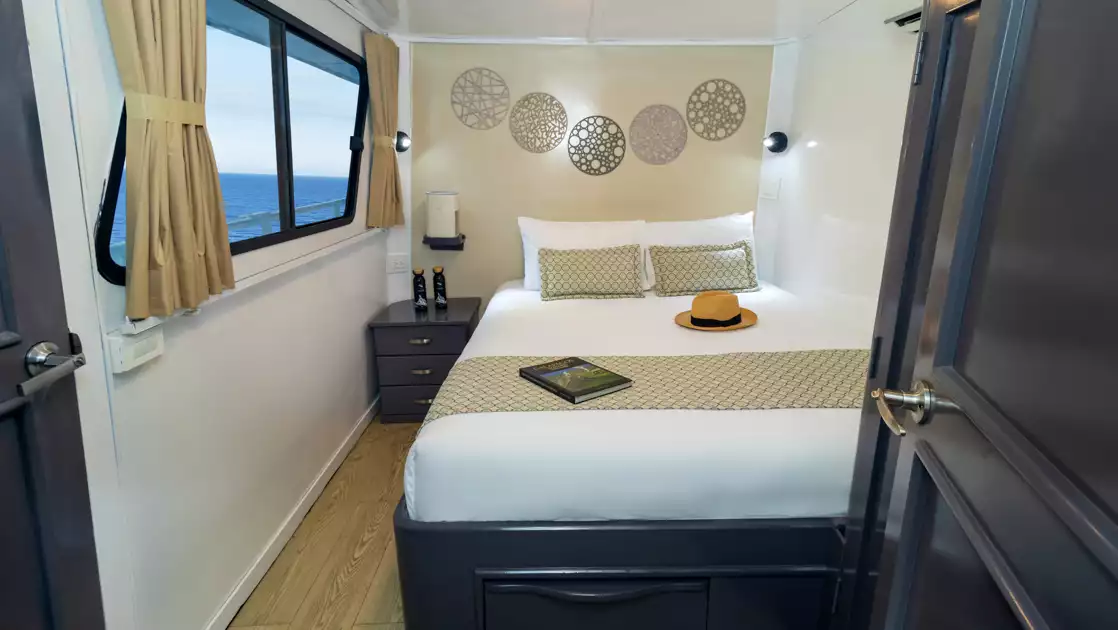 Upper Deck Cabin on Monserrat boat in Galapagos with matrimonial-size bed in white, beige accents & big view window.